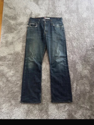 Blå Levi's 506 jeans med slitningar - Säljer ett par blå Levi's 506 jeans med raka ben och slitna detaljer vid fickorna. Jeansen har klassisk femficksmodell, normal midja och gul kontrastsöm. Perfekta för en avslappnad och trendig look.  Mått:  Ben utsida: 106 cm Midja: 43 cm Benöppning: 23 cm Ben insida: 78 cm