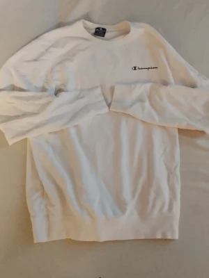Champion, Vit sweatshirt - Säljer en stilren vit sweatshirt från Champion med broderad logga på bröstet och en liten logga på ärmen. Tröjan har rund halsringning, långa ärmar och ribbade muddar. Perfekt för en clean och avslappnad look.