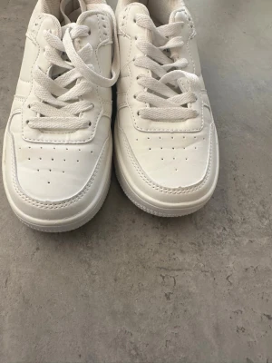 Vita sneakers med låg profil ! - Säljer ett par helvita sneakers med låg profil, breda vita skosnören och perforerade detaljer på tån. Skorna har en chunky sula och är tillverkade i syntetmaterial för en clean och modern look.