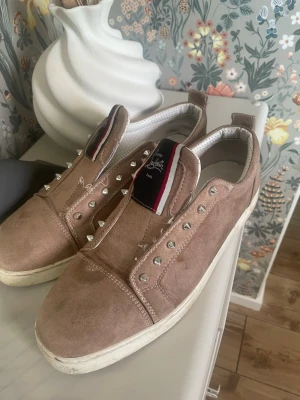Beige sneakers med nitar - Säljer ett par beige sneakers i mocka med silverfärgade nitar längs snörningen och vit sula. Skorna har en detaljrik plös med röd, vit och svart rand. Perfekta för dig som vill sticka ut med en edgy stil.