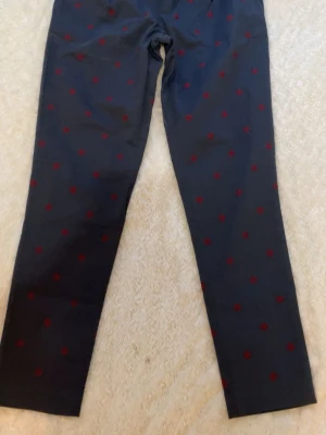 Mörkblå chinos med röda detaljer - Snygga mörkblå chinos från Tommy Hilfiger med röda broderade prickar över hela byxan. Klassisk rak passform och diskreta fickor. Tillverkade i mjukt bomullstyg för en bekväm känsla. Perfekta för dig som vill sticka ut lite extra.