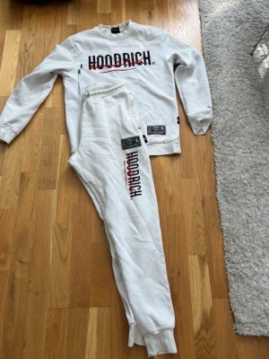 Vitt Hoodrich loungewear-set - Snyggt vitt loungewear-set från Hoodrich med matchande sweatshirt och byxor. Båda plaggen har Hoodrich-logga i svart och rött framtill samt coola patch-detaljer. Mjukt material och avslappnad passform, perfekt för chill dagar hemma.