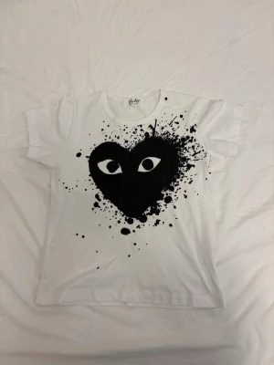 CDG Play tshirt - Vit t-shirt med ett stort svart hjärta och ögon på bröstet, omgiven av svart färgstänk. T-shirten har rund hals och korta ärmar. Perfekt för dig som gillar grafiska tryck och vill sticka ut lite extra.