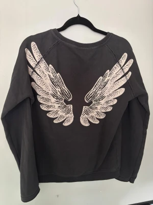 Svart Hunkydory sweatshirt med vingar - Cool svart sweatshirt med rund hals och långa ärmar. På ryggen finns ett stort tryck av vita änglavingar som ger tröjan en unik och edgy look. Perfekt för dig som vill sticka ut lite extra. Tillverkad i mjukt bomullsmaterial.