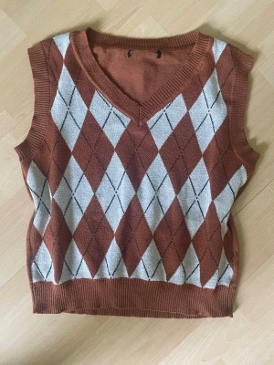 Stickad väst - Snygg stickad slipover i brunt med klassiskt argylemönster i vitt och svart på framsidan. Västen har v-ringning och ribbade kanter vid hals, ärmhål och nederkant. 