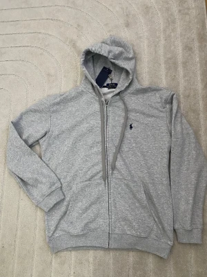 Grå zip hoodie från Polo Ralph Lauren L - Snygg grå hoodie med dragkedja från Polo Ralph Lauren. Klassisk design med huva och dragsnören, två fickor framtill och den ikoniska lilla loggan broderad på bröstet. Tillverkad i mjukt sweatshirtmaterial som är perfekt för chill dagar.