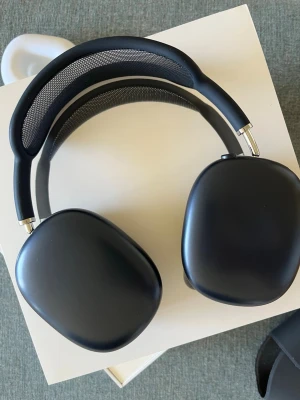 Svarta over-ear hörlurar - Stilrena svarta over-ear hörlurar med mjuka, utbytbara öronkuddar i tyg och en båge med mesh för extra komfort. Hörlurarna har en modern, rundad design och metalliska detaljer vid gångjärnen. Perfekta för musik, gaming eller film.