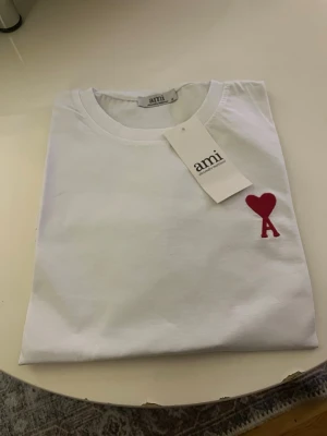 Vit t-shirt från Ami med rött hjärta( kom med bud) - Snygg vit t-shirt från Ami med ett rött hjärta och bokstaven A broderat på bröstet. Tillverkad i ekologisk bomull enligt GOTS-standard. Klassisk passform och rund halsringning, perfekt till alla dina outfits.