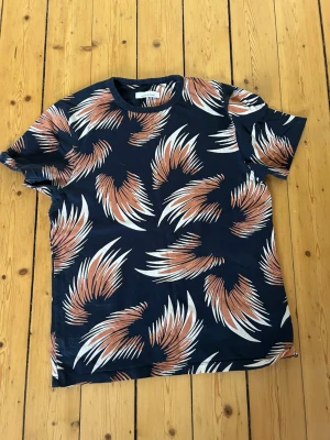 Mönstrad t-shirt från Samsøe Samsøe - Snygg t-shirt från Samsøe Samsøe med coolt abstrakt mönster i vitt och rostbrunt på mörkblå botten. Klassisk rund hals och korta ärmar. Perfekt för dig som vill sticka ut lite extra i vardagen.