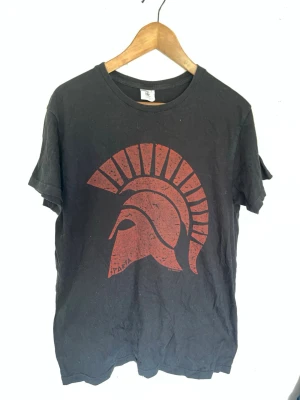Svart t-shirt med röd spartahjälm - Cool svart t-shirt med ett stort rött tryck av en spartansk hjälm på bröstet. Klassisk rund halsringning och korta ärmar. Perfekt för dig som gillar stilrena motiv och vill sticka ut lite extra.