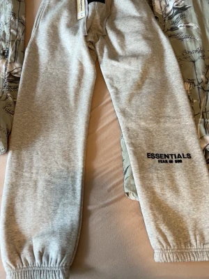 Gråa Essentials mjukisbyxor S - Snygga ljusgrå mjukisbyxor från Essentials Fear of God med resår i midjan och benslut. Byxorna har dragsko i midjan och logga tryckt på vänster ben. Perfekta för en chill och avslappnad stil. Priset kan diskuteras