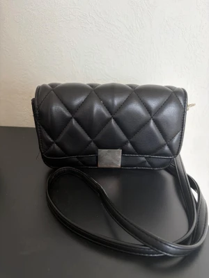 Svart axelväska  - Snygg svart axelväska med mönster. Perfekt för dig som vill ha en trendig och enkel accessoar till din outfit.