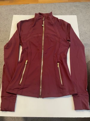 Vinröd träningsjacka från Lululemon - Snygg vinröd träningsjacka från Lululemon med figurnära passform, hög krage och dragkedja framtill. Två fickor med dragkedja och guldfärgade detaljer. Tillverkad i stretchigt funktionsmaterial som passar perfekt till träning eller som ett extra lager. Storlek 4