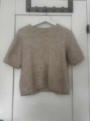 Beige stickad kortärmad tröja - Mysig beige stickad tröja med korta ärmar och rund hals. Tröjan har en rak, något boxig passform och är tillverkad i ett mjukt, lätt material som ger en avslappnad look. Perfekt att styla med jeans eller kjol för en enkel och trendig outfit. Stlk 158/164