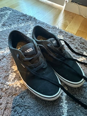 Svarta Vans sneakers med vita sulor - Säljer ett par klassiska svarta Vans sneakers med lågt skaft och vita gummisulor. Skorna har svart textilmaterial, svarta skosnören och Vans-logga på plösen och sidan. Perfekta för en avslappnad och stilren look.