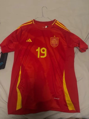 Spansk landslagströja röd gul - Säljer en röd fotbollströja från Spaniens landslag med gula detaljer och nummer 19 på framsidan. På ryggen står det 'LAMINE YAMAL' och nummer 19 i gult. Tröjan har korta ärmar, rund hals och är tillverkad i ett lätt funktionsmaterial.