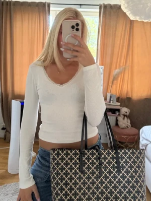 Vit långärmad topp med spetskant - Säljer en vit långärmad topp med v-ringning och fin spetskant runt halsen. Toppen är croppad och har en slim passform som sitter snyggt på kroppen. Bilden är lånad
