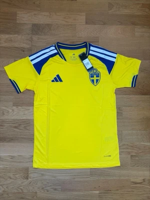 Svensk landslagströja Adidas gul - Gul svensk landslagströja från Adidas med blå detaljer och vita ränder på axlarna. Tröjan har broderat landslagsemblem på bröstet och klassisk Adidas-logga. Tillverkad i lätt och ventilerande material, perfekt för fotboll eller träning.
