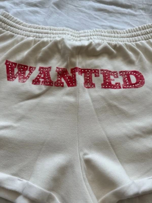 Vita shorts med röd text och cowboy-boot från Pink - Vita shorts med resår i midjan och snörning. På baksidan står det 'WANTED' i röd text med vita prickar och framtill finns en röd cowboy-boot med stjärnor. Mjukt material, perfekta för en avslappnad look.
