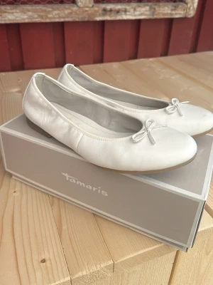 Vita ballerinaskor från Tamaris - Snygga vita ballerinaskor från Tamaris i mjukt skinn med rund tå och liten rosett framtill. Skorna har låg klack och klassisk design som passar till många olika outfits. Perfekta för dig som gillar stilrena och bekväma skor. Använda en gång, men säljes pga för små, nypris är 799kr