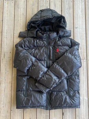Svart pufferjacka från Polo Ralph Lauren - OBS! SAKNAR ALLA LAPPAR ENDAST EN STORLEKSLAPP PÅ INSIDAN! Snygg svart pufferjacka från Polo Ralph Lauren med röd broderad logga på bröstet. Jackan har huva, dragkedja framtill och två sidofickor med dragkedja. Perfekt för kalla vinterdagar och riktigt skön att bära. Materialet är glansigt och quiltat för extra värme.