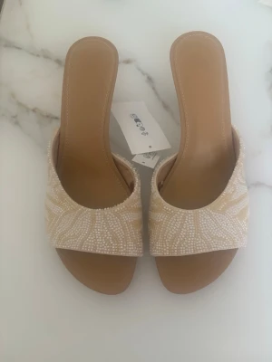 Beige sandaler med pärlbroderi - Säljer ett par snygga beige sandaler med öppen tå och bred rem dekorerad med vita pärlor i ett blommigt mönster. Sandalerna har en låg klack och är gjorda i konstläder med mjuk innersula för extra komfort. Perfekta för en trendig sommarlook.