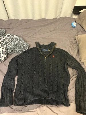 Svart kabelstickad tröja Polo Ralph Lauren quarter zip - Mörkgrå kabelstickad tröja från Polo Ralph Lauren med halv dragkedja och ståkrage. Tröjan har det klassiska röda Polo-logot på bröstet och ribbade muddar vid ärmslut och nederkant. Perfekt för lager på lager och snygg till jeans.