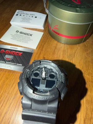 Svart Casio G-Shock klocka - Tuff svart Casio G-Shock klocka med robust design och digitalt-analogt urverk. Klockan har en bred rem i syntetmaterial och tydliga detaljer på urtavlan. Perfekt för dig som gillar sportig och modern stil. Originalförpackning och manual medföljer.