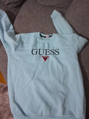 Ljusblå sweatshirt från Guess - Ljusblå sweatshirt från Guess med klassisk logga och röd triangel på bröstet. Tröjan har rund halsringning, långa ärmar och ribbade muddar vid ärmslut och nederkant. Perfekt för en avslappnad och trendig look.