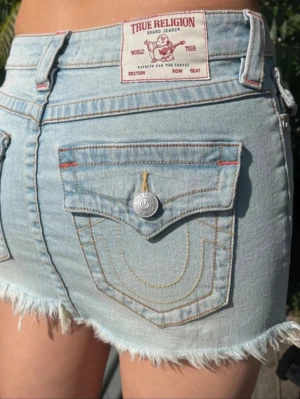 Ljusblå jeanskjol från True Religion - Trendig ljusblå jeanskjol från True Religion med rå kant nertill och klassiska bakfickor. Kjolen har en kort passform och är tillverkad i denim med snygga slitningar för en avslappnad look. Perfekt till sommaren och enkel att matcha med olika toppar.