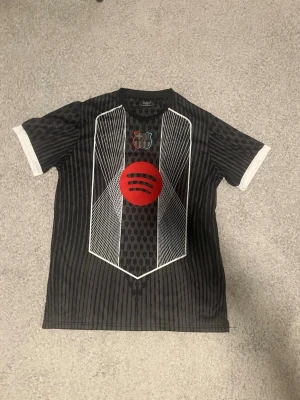 Svart FC Barcelona matchtröja - Säljer en svart FC Barcelona matchtröja med vita och röda detaljer. Tröjan har ett stort rött Spotify-tryck på bröstet, vita grafiska linjer och klubbens emblem i svart, rött och blått. Korta ärmar med vita muddar och sportig design. Perfekt för fotbollsträning eller support. står xxl men passar som xl
