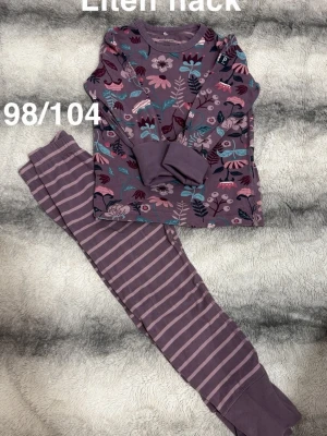 Lila blommig pyjamas med ränder - Superfin tvådelad pyjamas i lila toner. Tröjan har ett färgglatt blommönster, medan byxorna är randiga i två nyanser av lila. perfekt för mysiga kvällar hemma.🌸 Liten fläck. Storlek 98/104