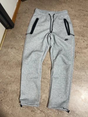 Gråa mjukisbyxor från Nike - Snygga ljusgrå mjukisbyxor från Nike med svarta detaljer, dragsko i midjan och dragkedjor på fickorna. Perfekta för chill eller träning. Klassisk Nike-logga på benet och smal passform med muddar vid benslut.