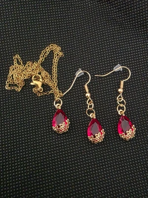 Smyckeset med röda droppar - Snyggt smyckeset med halsband och örhängen i guldton med röda droppformade stenar. Stenarna har en glittrande yta och dekoreras med små guldfärgade detaljer längst ner. Perfekt för att lyfta vilken outfit som helst.