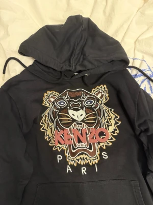 Svart Kenzo hoodie med tigertryck - Svart hoodie från Kenzo med ikoniskt broderat tigerhuvud och texten 'KENZO PARIS' på bröstet. Hoodien har huva med dragsko och en stor magficka. Perfekt för dig som vill sticka ut med en streetstyle-look.