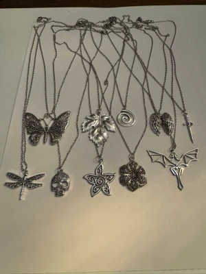 10 halsband med unika hängen i silverfärg - Säljer ett paket med 10 halsband i silverfärg, alla med olika hängen som fjäril, trollslända, dödskalle, löv, spiral, stjärna, blomma, fladdermus, vingar och kors. Kedjorna är tunna och smidiga, och hängena har coola detaljerade mönster. Perfekt för dig som gillar alternativ stil. Smyckena sälja även separat i andra annonser .  Mestadels är materialet i smyckena zinklegering, kedja rostfritt vilket iaf 1 av smyckena är med