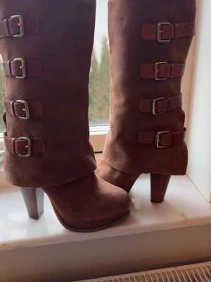 Bruna stövletter med spännen och klack - Snygga bruna stövletter i mockaimitation med höga klackar och flera dekorativa spännen i guld längs skaftet. Stövletterna har rundad tå och grov klack, perfekt för dig som vill sticka ut med din stil.