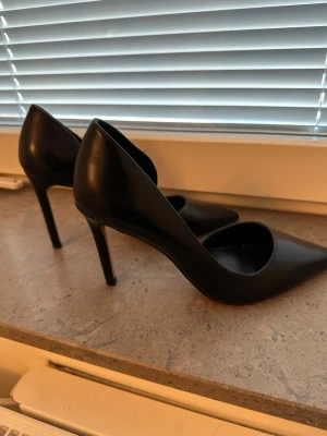 Svarta klassiska pumps med spetsig tå - Stilrena svarta pumps med spetsig tå och hög klack. Skorna har en elegant och tidlös design som passar perfekt till många olika outfits. Tillverkade i ett slätt material med en klassisk siluett och öppen insida för en modern touch.