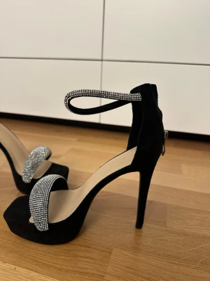 Svarta pumps med strassdetaljer - Snygga svarta pumps med höga smala klackar och platåsula. Skorna har en bred rem över tårna och en smal ankelrem, båda täckta av glittrande strass. Materialet är mockaimitation och insidan är ljusbeige. Perfekta för att sticka ut.