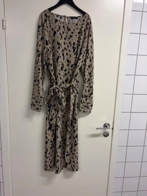 Beige långklänning från Rut&Circle - Ny oanvänd klänning. Snygg beige långklänning från Rut&Circle med svart leopardmönster. Klänningen har långa ärmar, rund halsringning och ett matchande knytbälte i midjan. Perfekt för dig som gillar trendiga och bekväma plagg med coolt mönster. Upplever den som mindre i storleken mer strl L 