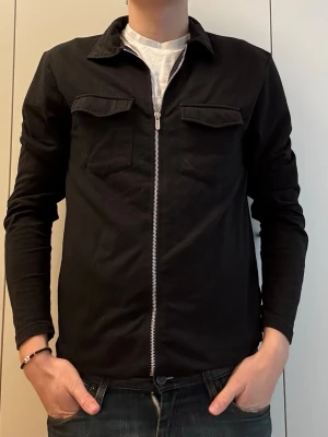 Svart overshirt med dragkedja MAN - Stilren svart overshirt från MAN med dragkedja framtill och två bröstfickor med lock. Overshirten har klassisk krage och raka ärmar, perfekt för lager på lager. Materialet känns mjukt och bekvämt, passar till många olika looks. Längd: 70cm, P2P: 48cm