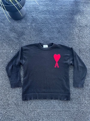 Ami Paris stickad tröja - Snygg svart stickad tröja från Ami paris med röd hjärtlogga och bokstaven A på bröstet. Tröjan har rund halsringning, ribbade muddar och nederkant. Perfekt för dig som gillar stilrena plagg med en twist. Storlek M, skick 8/10. Pris går att diskutera✅
