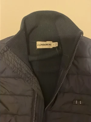 Mörkblå pufferjacka från J.Lindeberg - Snygg mörkblå pufferjacka från J.Lindeberg med quiltad framsida och stickade ärmar. Jackan har dragkedja framtill och diskret logga på bröstet. Perfekt för kalla dagar och ger en sportig look. Ny pris 4000 kr