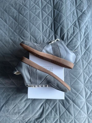 Grå sneakers från Maison Margiela - Snygga sneakers i grå mocka och läder från Maison Margiela. Skorna har vita snören, beige innersula och klassisk gummisula i brunt. Designen är stilren med kontrasterande material och diskreta detaljer. Perfekta för dig som gillar minimalistisk och modern stil.