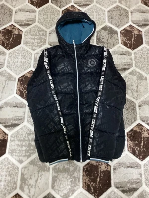 Svart pufferjacka från Salty Dog - Snygg svart pufferjacka från Salty Dog med tryckt logga på bröstet och textdetaljer över hela jackan. Jackan har huva med blått innerfoder och vita dragkedjor samt breda band med text längs dragkedjan. Perfekt för kalla vinterdagar.