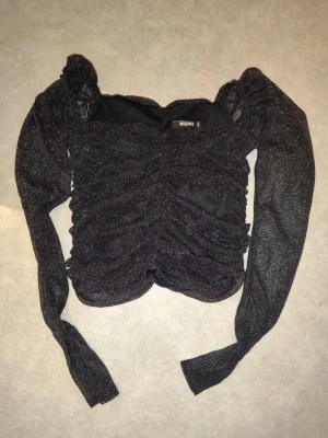 Glittrig svart topp party - Säljer en svart glittrig topp med rynkade detaljer.. Toppen har en croppad passform och är perfekt för dig som vill sticka ut lite extra. Materialet är glittrigt och toppen har en snygg offshoulder-design.