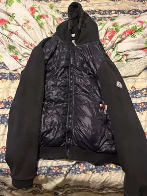Svart hybridjacka med huva från Moncler - Snygg svart hybridjacka från Moncler med quiltad, glansig front i polyester och mjuka ärmar samt rygg. Jackan har huva, dragkedja framtill och Moncler-logga på ärmen. Perfekt för dig som vill ha en sportig och stilren look.
