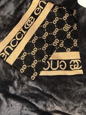 Beige och svart halsduk från Gucci - Snygg halsduk från Gucci i beige och svart med klassiskt GG-mönster och stora loggor längs kanterna. Halsduken är rektangulär och har en lyxig känsla, perfekt för att lyfta vilken outfit som helst. Materialet känns mjukt och skönt mot huden.