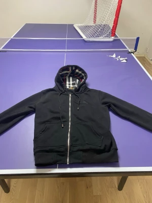 Svart Burberry hoodie med dragkedja - Svart hoodie från Burberry med dragkedja framtill och klassiskt rutigt foder i huvan. Broderad logga på bröstet och stora fickor framtill. Perfekt för en stilren och avslappnad look. Materialet är mjukt och bekvämt, troligen bomullsblandning.