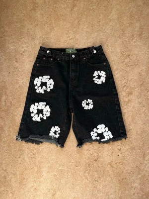Svarta jeansshorts med blommönster - Unika svarta jeansshorts från Second Phase med vita blommiga tryck över hela plagget. Shortsens ben är råkantade för en avslappnad look och de har klassisk femficksdesign. Perfekta för dig som vill sticka ut med en kreativ och trendig stil.